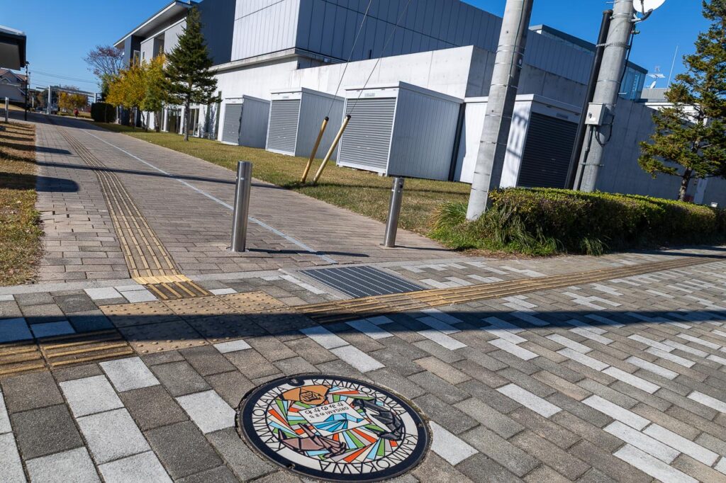 上三川町A「ORGAMI」のデザインマンホール蓋と設置場所の周囲がわかる現地の様子